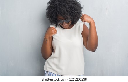 Young african american plus size woman over grey grunge wall wearing fashion sunglasses very happy and excited doing winner gesture with arms raised, smiling and screaming for success. Celebration