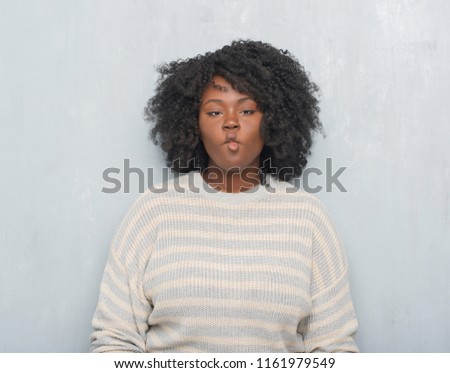 Young african american plus size woman over grey grunge wall wearing a sweater making fish face with lips, crazy and comical gesture. Funny expression.