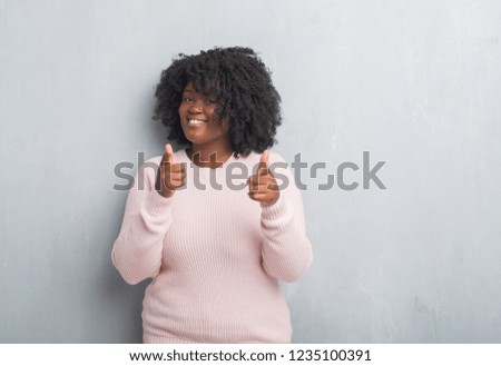 Young african american plus size woman over grey grunge wall wearing winter sweater pointing fingers to camera with happy and funny face. Good energy and vibes.