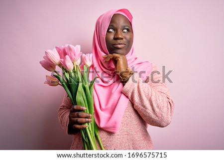 Young african american plus size woman wearing muslim hijab holding bouquet of pink tulips with hand on chin thinking about question, pensive expression. Smiling with thoughtful face. Doubt concept.