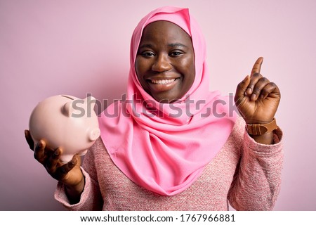 Young african american plus size woman wearing muslim hijab holding pink piggy bank surprised with an idea or question pointing finger with happy face, number one
