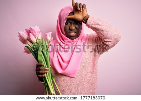 Young african american plus size woman wearing muslim hijab holding bouquet of pink tulips making fun of people with fingers on forehead doing loser gesture mocking and insulting.