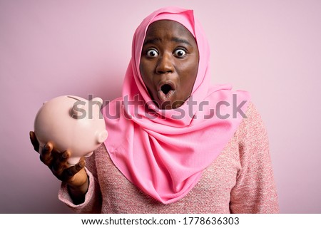 Young african american plus size woman wearing muslim hijab holding pink piggy bank scared in shock with a surprise face, afraid and excited with fear expression