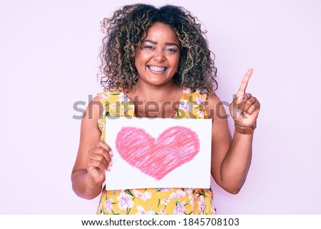 Young african american plus size woman holding heart shape paper surprised with an idea or question pointing finger with happy face, number one 