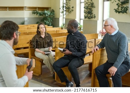 Young African American priest with Bible talking to one of parishioners while explaining her some verses from New Testament
