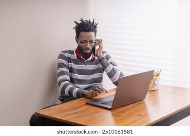 Young African American professional in casual striped sweater working from home. Man with dreadlocks and glasses using laptop and smartphone for business call, showcasing modern remote work lifestyle. - Powered by Shutterstock