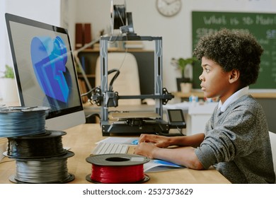 Young African American pupil inspecting design on computer in a modern classroom filled with technology and 3D printing equipment without looking at the camera