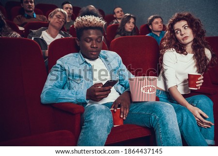 Young African American rewrites in chat using phone. A woman looks at a man who is looking at the phone while sitting in the cinema. 
