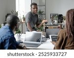 Young African American an sitting on white table while telling joke during conversation with his coworkers in office