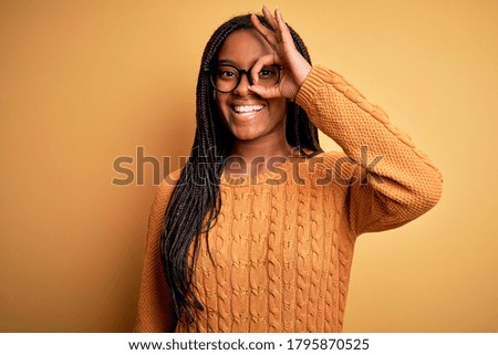 Young african american smart woman wearing glasses and casual sweater over yellow background doing ok gesture with hand smiling, eye looking through fingers with happy face.
