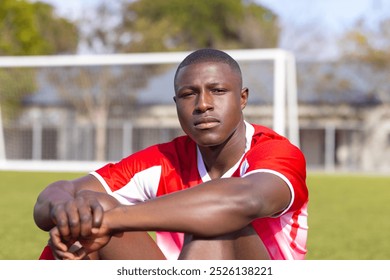 Young African American soccer player sitting on soccer field, looking focused. Green grass and goalposts in background, with trees and buildings visible, unaltered - Powered by Shutterstock