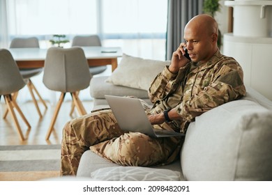Young African American soldier communicating over cell phone while surfing the net on laptop at home.