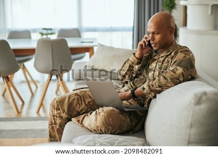 Young African American soldier communicating over cell phone while surfing the net on laptop at home.