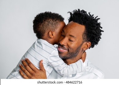 A young African American son hugs his father around the neck.  They are both dressed in white against a white background.