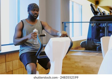 Young African American sportsman looking at monitor while he running exercise bike, he drinking pure water to control water balance - Powered by Shutterstock