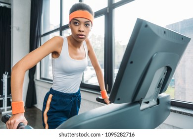 Young African American Sportswoman In Headband And Wristbands Running On Treadmill At Gym 