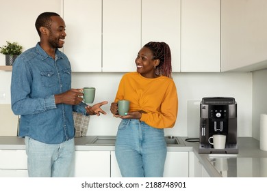 Young African American Spouses Drinking Morning Coffee And Talking While Standing Near Coffee Machine In Kitchen Interior. Black Couple Enjoying Time At Home