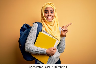 Young African American Student Girl Wearing Muslim Hijab And Backpack Holding Notebook Very Happy Pointing With Hand And Finger To The Side