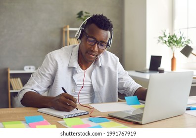 Young african american student in headset studying online writing, listening and watching webinar, training course using laptop computer and notebook for making notes. Remote education, e-learning