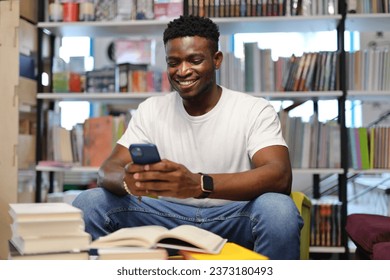 A young African American student in a modern library, studying with a cellphone and books, pursuing education happily.