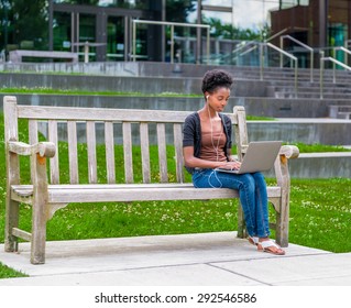 Young African American Student Sitting On Bench Doing Homework While Listening To Music