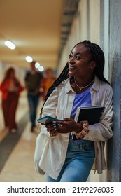 A Young African American Student Is Standing In The Corridor At The University With A Smartphone In Her Hand. She Is Waiting For Her Next Class And Is Killing Time By Checking Her Social Media Online.