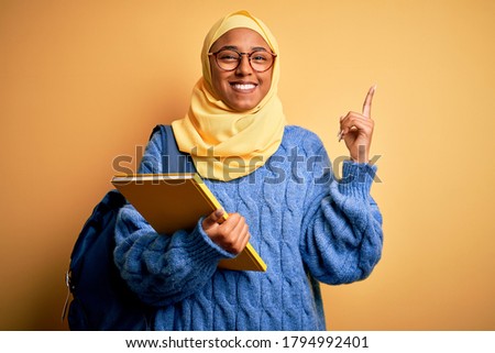 Young African American student woman wearing muslim hijab and backpack holding book with a big smile on face, pointing with hand finger to the side looking at the camera.