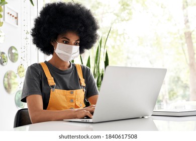 Young African American Teen Girl Student With Afro Hair Wearing Face Mask Using Laptop Sitting At Cozy Cafe Table Alone Indoor. Social Distancing And Work, Study On Computer With Covid 19 Protection.