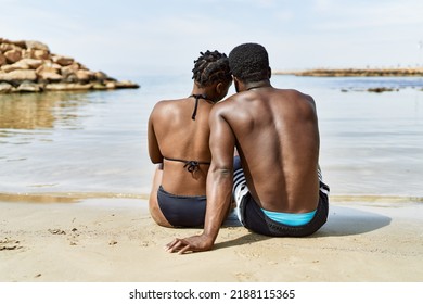 Young african american tourist couple on back view wearing swimwear sitting at the beach. - Powered by Shutterstock