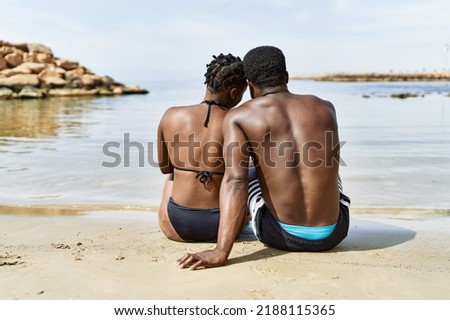 Young african american tourist couple on back view wearing swimwear sitting at the beach.