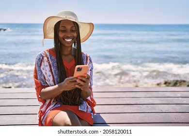 Young african american tourist woman using smartphone sitting on the bench at the beach.