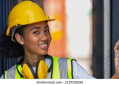 Young African American trainee in safety suit, yellow hard hat and earmuffmuff stand smiling next to big container in a shipyard. African curly hair female worker in smile. Female loader with earmuff - Shutterstock ID 2129328284