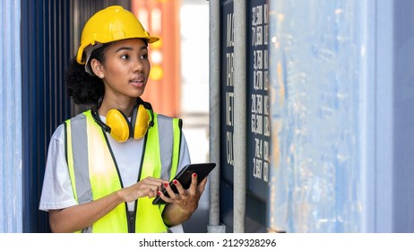 Young African American trainee in safety suit, yellow hard hat and earmuff uses tablet or smart phone in a shipyard. African curly hair female worker in smile. Female loader with yellow earmuff - Powered by Shutterstock