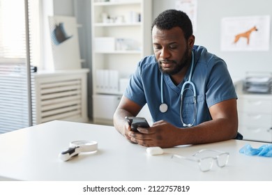 Young African American Veterinarian In Blue Uniform Scrolling In Mobile Phone While Sitting By Workplace In Hospital