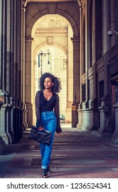 Young African American Woman with afro hairstyle, wearing mesh sheer long sleeve shirt blouse, blue jeans, sandals, holding leather folder, looking around, walking on narrow vintage street in New York