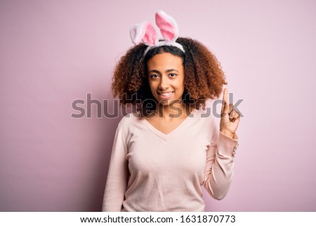 Young african american woman with afro hair wearing bunny ears over pink background pointing finger up with successful idea. Exited and happy. Number one.