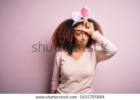 Young african american woman with afro hair wearing bunny ears over pink background making fun of people with fingers on forehead doing loser gesture mocking and insulting.
