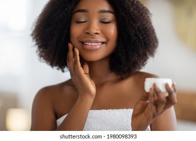 Young African American Woman Applying Hydrating Cream On Her Face, Smiling With Closed Eyes, Indoors. Millennial Black Female Making Daily Domestic Skincare Routine, Using Cosmetic Product