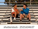 A young African American woman and an Asian man, both athletes, sit on bleachers, smiling and fist-bumping. The athletes enjoy a break on the bleachers after workout. Diverse athletic couple on bench