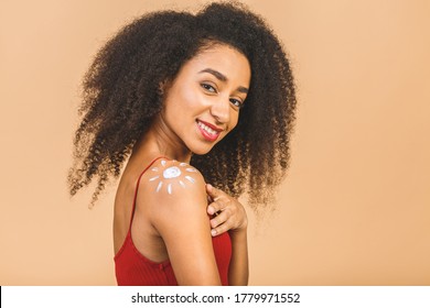 Young african american woman with bare shoulders standing isolated on beige background applying cream on body pointing at sun shape smiling joyful. 
