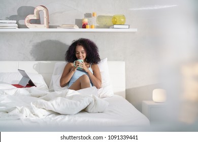 Young African American woman in bed at home during weekend. Happy black girl smiling while watching movie on laptop computer and drinking coffee - Powered by Shutterstock