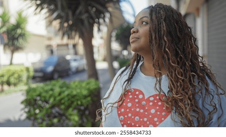 Young african american woman with braided hair wearing a heart-patterned shirt, standing on an urban street, looking thoughtful and confident in a city environment. - Powered by Shutterstock