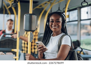 Young african american woman with braids smiling while standing by herself on a bus and listening to music on a smartphone. Happy female passenger in public transportation. Looking into the camera. - Powered by Shutterstock