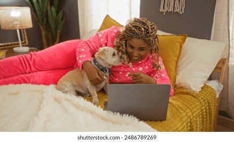 Young african american woman with braids wearing pink pajamas on a bed in a cozy bedroom, using a laptop while cuddling her small dog. - Powered by Shutterstock