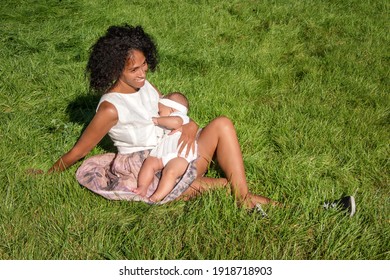 Young african american woman breastfeeding her baby sitting on grass in summer park. Infancy, motherhood, lactation, nutrition and outdoors breastfeeding concept. Black mother breast feeding her child