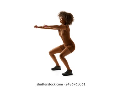 Young African American woman in brown activewear doing squats lifting small lifting small weights against white studio background. Concept of sport, mourning routine, active lifestyle, energy, action.