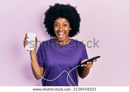 Young african american woman charging smartphone using portable battery celebrating crazy and amazed for success with open eyes screaming excited. 