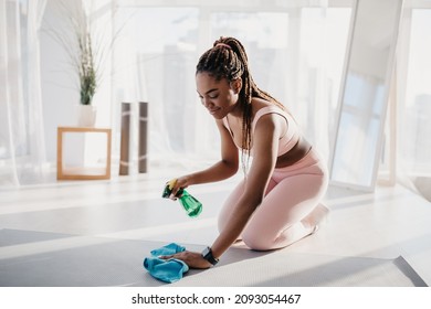 Young African American Woman Cleaning Yoga Mat After Workout At Fitness Studio, Copy Space. Millennial Black Lady Using Detergent Spray, Wiping Sports Equipment In Domestic Gym