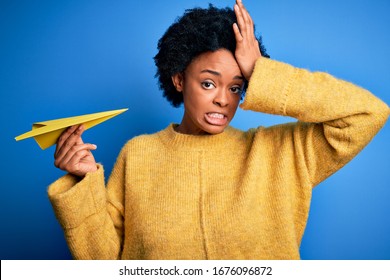 Young African American Woman With Curly Hair Holding Paper Airplane Over Blue Background Stressed With Hand On Head, Shocked With Shame And Surprise Face, Angry And Frustrated. Fear And Upset 