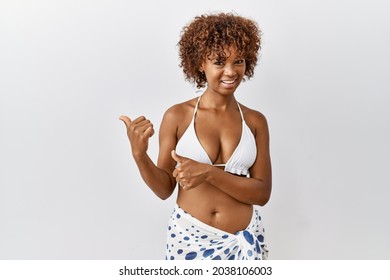 Young african american woman with curly hair wearing bikini pointing to the back behind with hand and thumbs up, smiling confident 
